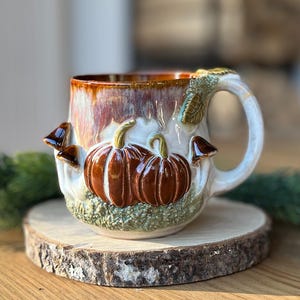 Peut inclure: Une tasse en céramique avec une finition émaillée brillante blanche et brune. L'extérieur est orné de deux citrouilles orange en relief, de plusieurs petits champignons bruns et d'une base verte texturée. Une anse est fixée sur le côté.