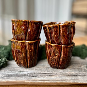 Puede incluir: Cuatro tazas de cerámica hechas a mano con un diseño texturizado similar a la corteza. Las tazas son de un color marrón oscuro intenso con un acabado brillante. Las tazas están apiladas en pares, mostrando su forma única.