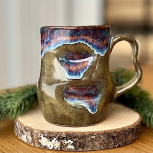 Pode incluir: Uma caneca de cerâmica artesanal com uma vidragem única. A caneca apresenta uma base castanha com detalhes azuis, roxos e brancos. Possui uma alça curva e assenta numa base de madeira. A caneca tem aproximadamente 10 cm de altura.