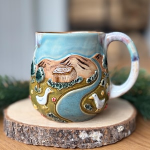 Può includere: Una tazza in ceramica con un paesaggio in rilievo dipinto a mano che raffigura una piccola capanna, colline marroni, un fiume blu tortuoso e due anatre bianche. La tazza presenta un cielo azzurro, un primo piano verde e un manico curvo.