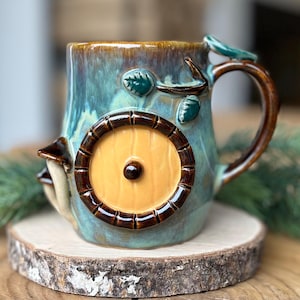 Peut inclure: Une tasse en céramique au design fantaisiste, avec une porte jaune ronde encadrée de marron et un bouton marron foncé. La tasse a un glaçage bleu-vert et marron, avec une anse en forme de branche et des feuilles décoratives.