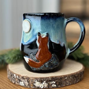 May include: A ceramic mug featuring a glossy glaze in shades of deep black and light blue. A sculpted, three-dimensional orange fox sits on the front, facing a raised white circular moon. The mug has a curved handle and a smooth, rounded body.