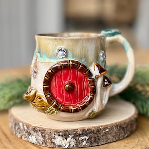 Può includere: Una tazza in ceramica dal design stravagante, con una porta rossa, funghi e decorazioni floreali. La tazza ha una combinazione di colori beige e verde e poggia su una fetta di legno. Il manico è verde.