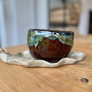 Op de afbeelding: Een keramische kom met een donkerbruine basis en een glanzende, groen uitgelopen glazuurrand, voorzien van een spiraalpatroon aan de zijkant. De kom staat op een lichtgekleurd, golvend keramisch schoteltje in de vorm van een blad met een steeltje.