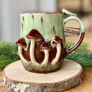 Może przedstawiać: Ceramiczny kubek z jasnozieloną i brązową glazurą, ozdobiony czterema brązowo-kremowymi rzeźbami grzybów. Uchwyt jest brązowy i zielony. Kubek stoi na drewnianym plastrze.