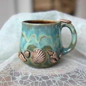 Puede incluir: Una taza de cerámica con un esmalte brillante en tonos azul verdoso, azul y verde oliva. El exterior está decorado con varias esculturas de conchas marinas tridimensionales en relieve con acabado en bronce metálico, incluida una vieira central.