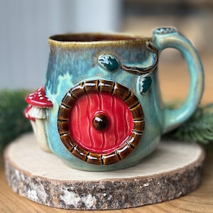 May include: A ceramic mug with a glossy blue-green glaze featuring a raised, circular red door with a brown frame and a small red mushroom with white spots on the side. The handle is shaped like a curved branch with small green leaves attached.