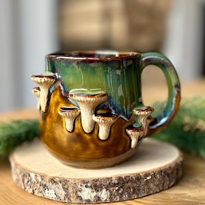 Puede incluir: Una taza de cerámica con acabado brillante, que presenta una base de color marrón oscuro que se transforma en un esmalte verde vibrante en la parte superior. El exterior está decorado con varios hongos esculpidos en 3D de color crema.