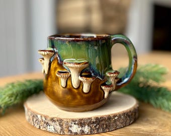Pode incluir: Uma caneca de cerâmica com acabamento brilhante, apresentando uma base castanho-escura que transita para um esmalte verde vibrante no topo. O exterior é decorado com vários cogumelos esculpidos em três dimensões na cor creme.