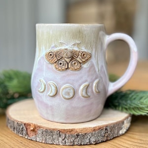 May include: A ceramic mug with a two-tone glaze featuring a cream-coloured top and a soft lavender bottom. The mug displays a raised, sculpted moth emblem centred above a horizontal row of embossed moon phases, including crescents and a full circle.