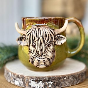 Puede incluir: Una taza de cerámica con la cara esculpida de una vaca de las Tierras Altas. La taza tiene una base verde, un borde marrón y un asa curvada. La cara de la vaca es de color canela con pelo y cuernos detallados. La taza se exhibe sobre una rodaja de madera.