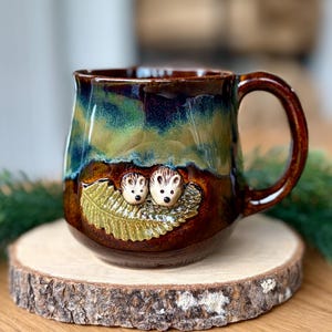 Könnte beinhalten: Ein Keramikbecher mit glänzender Glasur, der eine dunkelbraune Basis hat, die in einen leuchtend blauen und grünen oberen Bereich übergeht. Die Vorderseite zeigt ein erhabenes Relief von zwei kleinen Igeln auf einem grünen Farnblatt.