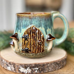 May include: A ceramic mug with a whimsical fairy door design. The mug features a teal and brown glaze, with a detailed door and mushroom accents. It sits on a wooden slice, with a green sprig in the background.