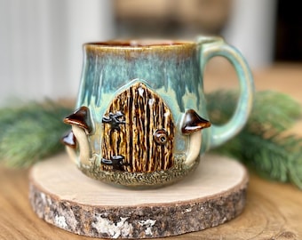 May include: A ceramic mug with a whimsical fairy door design. The mug features a teal and brown glaze, with a detailed door and mushroom accents. It sits on a wooden slice, with a green sprig in the background.