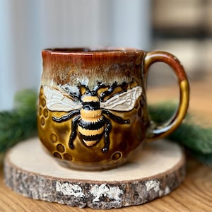 May include: A ceramic mug with a detailed bumblebee design. The mug has a brown and gold glaze, with a raised relief of a bee on the front. The handle and rim are also gold. The mug is on a wooden coaster.