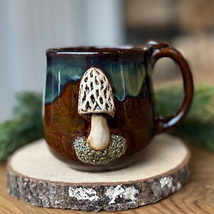 Può includere: Una tazza in ceramica fatta a mano con una glassa marrone, verde e blu. Una scultura dettagliata di un fungo è attaccata alla parte anteriore. La tazza è appoggiata su una fetta di legno, mostrando un'estetica rustica e terrosa.
