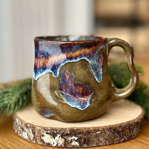 May include: A handmade ceramic mug with a unique glaze. The mug features a brown base with streaks of blue, purple, and orange. The handle is curved, and the mug sits on a small wooden slice. The mug is approximately 4 inches tall.