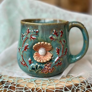 Puede incluir: Una taza de cerámica con esmalte azul verdoso brillante que presenta un relieve tridimensional de una concha abierta de color tostado con una perla blanca. La taza está decorada con algas de color marrón rojizo y texturas de coral.