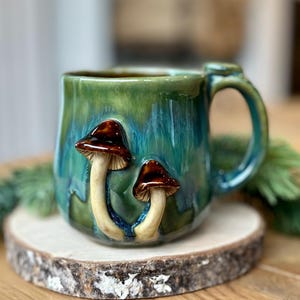 Puede incluir: Taza de cerámica con esmalte verde y azul, con dos acentos de hongos marrones y crema. La taza tiene un asa grande y se apoya en una rodaja de madera. Ideal para té o café.