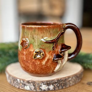 Könnte beinhalten: Ein Keramikbecher mit einer glänzenden Glasur in erdigen Braun- und Salbeigrüntönen. Die Außenseite zeigt dreidimensionale skulpturale Details, darunter kleine Blätter und einen Ast mit Pilzen. Der Becher hat einen dunkelbraunen Henkel.