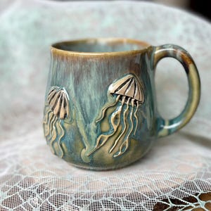 Könnte beinhalten: Eine Keramik-Tasse mit blau-grüner Glasur, die zwei erhabene Quallen-Designs in metallischer Goldfarbe aufweist. Die Tasse hat eine abgerundete Form mit Henkel und goldenem Rand.