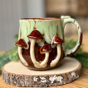 May include: A ceramic mug with a light green glaze and brown accents, adorned with four mushroom sculptures. The mushrooms have brown caps and white stems. The mug sits on a wooden slice.