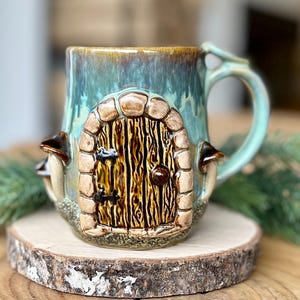 May include: A ceramic mug with a whimsical fairy door design. The mug features a teal and brown glaze, a door-shaped facade, and mushroom accents. The handle is a light teal color. The mug is placed on a wooden slice.