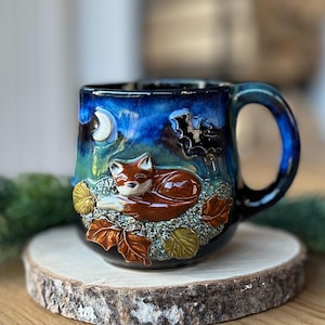 May include: A ceramic mug featuring a glossy deep blue and green glaze with a raised, sculpted scene of a sleeping orange fox resting on textured ground among brown and yellow leaves, under a white crescent moon and a black bat silhouette.