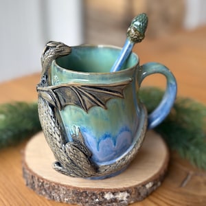 May include: A ceramic mug with a blue and green glaze featuring a sculpted silver dragon wrapped around the exterior. The mug includes a matching spoon with a textured, scale-like handle that rests inside the vessel, showcasing detailed craftsmanship.