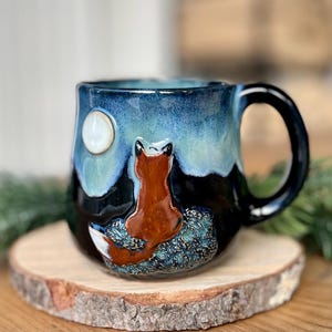 Peut inclure: Une tasse en céramique avec un motif de renard. La tasse présente une silhouette de renard brun contre un ciel nocturne avec une pleine lune. La tasse a un glaçage bleu foncé et noir avec une anse. La tasse est posée sur un dessous de verre en bois.