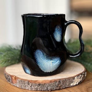 Pode incluir: Uma caneca de cerâmica preta com uma forma orgânica única e uma alça curva. A caneca apresenta detalhes em azul e branco e assenta numa base de madeira. É um artigo feito à mão.