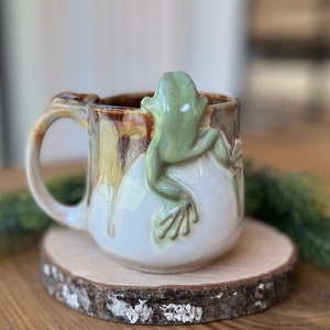May include: A ceramic mug featuring a light green frog climbing on the side. The mug has a white base with brown and yellow glaze. The handle and rim are brown. The mug sits on a wooden coaster.