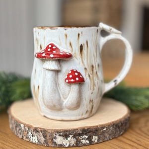 Può includere: Tazza in ceramica bianca con una glassa marrone e due accenti di fungo rosso e bianco. La tazza ha un manico ed è appoggiata su una fetta di legno.