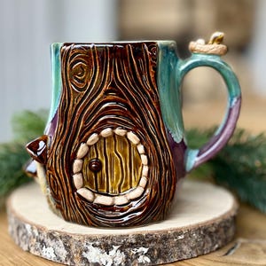 Peut inclure: Une tasse en céramique présentant un motif de tronc d'arbre brun texturé avec une porte circulaire et un petit champignon sur le côté. La poignée est ornée d'un petit détail sculpté au sommet, et la tasse a une finition brillante avec un émail bleu sarcelle et violet.
