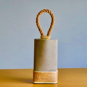 May include: A gray doorstop with a rope handle and a brown jute trim. The doorstop is sitting on a wooden surface.