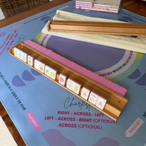 May include: A Mahjong set featuring pink and white tiles, wooden racks, and a blue and purple game mat. The mat displays the word "Charlest" and game instructions. A clear box contains extra tiles.