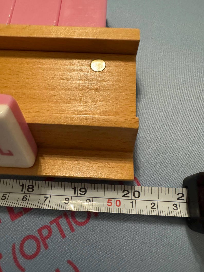 May include: Wooden game board with a light brown finish, featuring a recessed area and a small, round, gold-coloured accent. A measuring tape shows the board is approximately 50 cm long. Pink game pieces are visible.