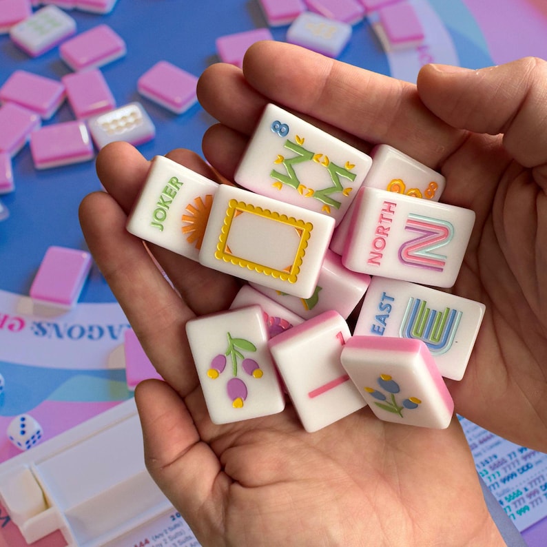 Może przedstawiać: Zbi&oacute;r białych i r&oacute;żowych kafelk&oacute;w do gry w Mahjong z r&oacute;żnymi kolorowymi symbolami i tekstem, w tym "JOKER", "NORTH" i "EAST". Kafelki są trzymane w dłoni, a dodatkowe kafelki są rozrzucone w tle.