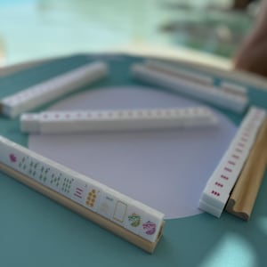 May include: A set of white mahjong tiles with pink dots and various symbols on a teal surface. The tiles are arranged in a circle with one tile set apart. The tile set apart has a wooden base and features a dragon, wind, and bamboo symbols.