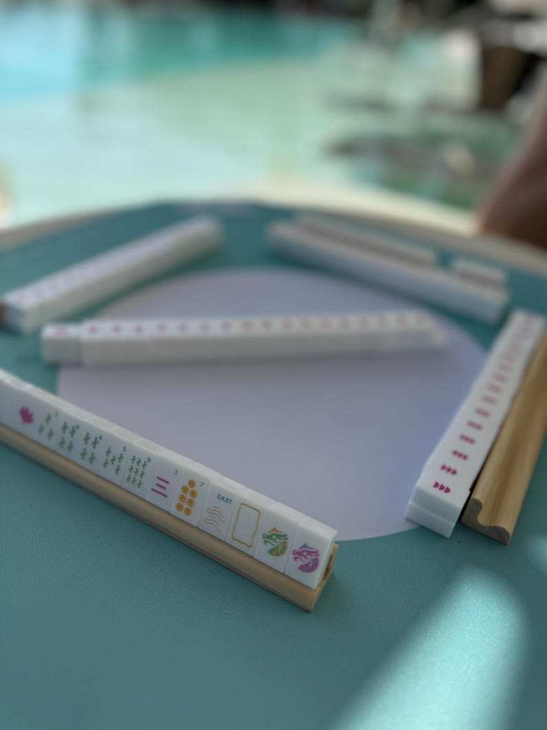 May include: A set of white mahjong tiles with pink dots and various symbols on a teal surface. The tiles are arranged in a circle with one tile set apart. The tile set apart has a wooden base and features a dragon, wind, and bamboo symbols.