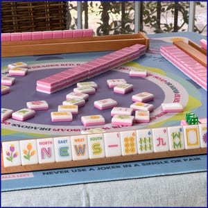 May include: A Mahjong set with pink and white tiles on a blue playing surface. The tiles, with various symbols, are arranged near wooden racks. The set includes green dice. The game is set up for play.