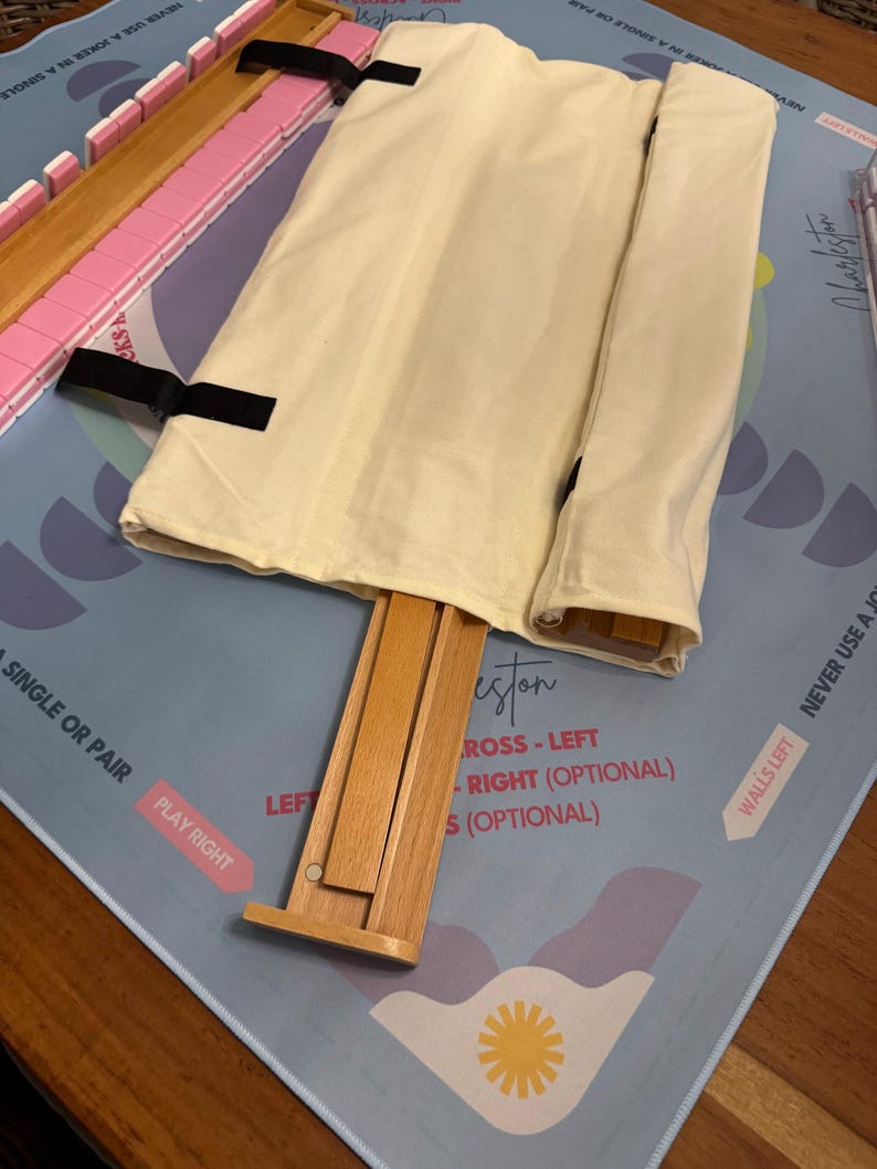 May include: A light blue game mat with text and illustrations, featuring a wooden rack and pink tiles. A beige cloth bag is secured with black straps. The game appears to be a version of Mahjong.