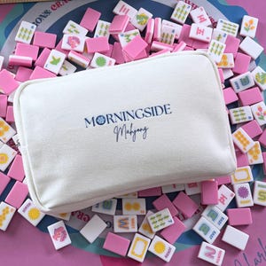 Może przedstawiać: Biała pł&oacute;cienna torba z napisem "MORNINGSIDE Mahjong" leży wśr&oacute;d rozrzuconej kolekcji r&oacute;żowych i białych płytek do mahjonga. Płytki przedstawiają r&oacute;żne symbole i znaki, tworząc kolorową i wciągającą ekspozycję.