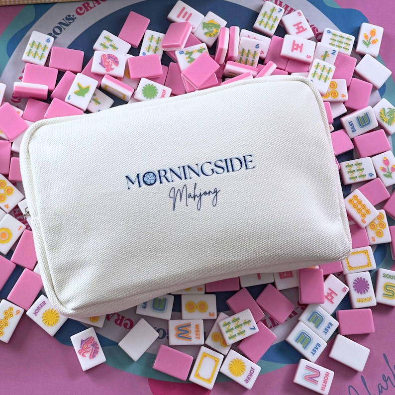 Może przedstawiać: Biała pł&oacute;cienna torba z napisem "MORNINGSIDE Mahjong" leży wśr&oacute;d rozrzuconej kolekcji r&oacute;żowych i białych płytek do mahjonga. Płytki przedstawiają r&oacute;żne symbole i znaki, tworząc kolorową i wciągającą ekspozycję.