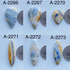 May include: Six polished gemstones labeled A-2266, A-2267, A-2270, A-2271, A-2272, and A-2273. Each stone features unique swirling patterns of blue, white, and yellow-brown colors. A silver-colored coin is placed next to each stone for scale.