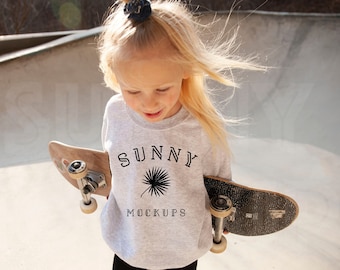 Może przedstawiać: Młoda osoba w szarej bluzie z napisem "SUNNY MOCKUPS" i grafiką palmy. Trzyma dwie deski skateboardowe.