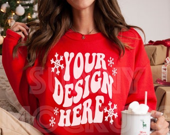May include: A red sweatshirt featuring a white snowflake design and the text "YOUR DESIGN HERE" in a retro font. The model is wearing the sweatshirt and holding a mug of hot chocolate with marshmallows.