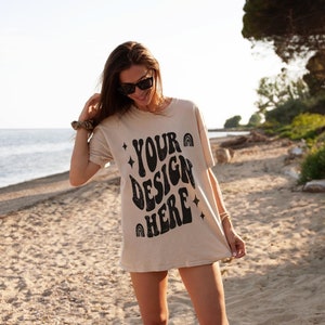 Puede incluir: Una mujer lleva una camiseta beige con el texto "YOUR DESIGN HERE" impreso en negro. La camiseta tiene un estilo de fuente retro y groovy. Est&aacute; de pie en una playa de arena con un oc&eacute;ano azul de fondo.