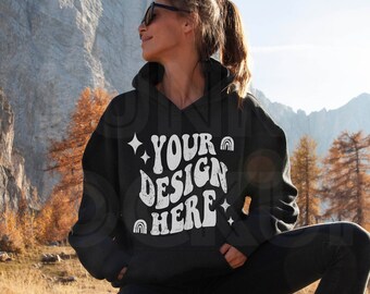 Puede incluir: Una sudadera con capucha negra con el texto "YOUR DESIGN HERE" en blanco, con un arcoíris y estrellas. La sudadera la lleva puesta una persona de pie en un paisaje montañoso.