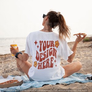 May include: A white t-shirt with the text "YOUR DESIGN HERE" in a retro font with red and orange accents and a flower graphic. The shirt is being worn by a person sitting on a beach.